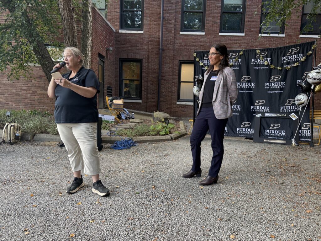 Speaker delivers remarks while another presenter stands nearby during an outdoor Purdue College of Agriculture welcome event.