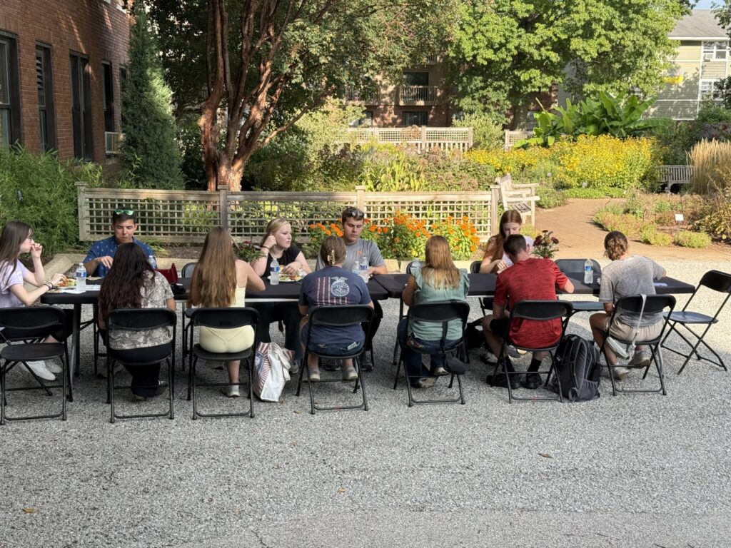Students seated together at an outdoor dinner surrounded by flowering garden beds and landscape plantings.
