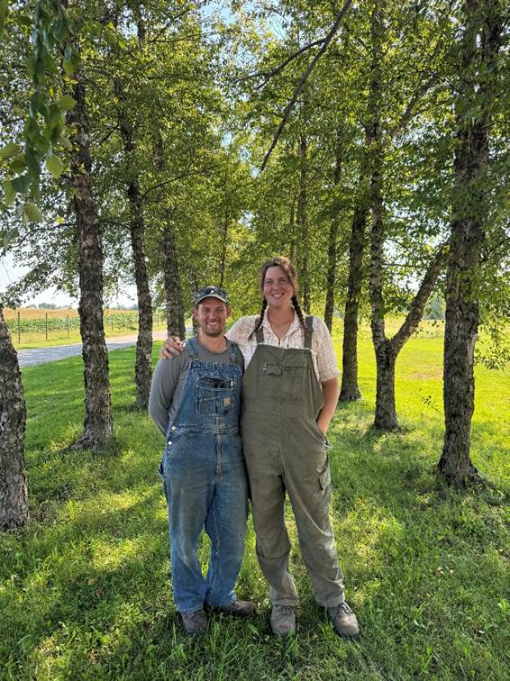 Two individuals wearing overalls stand together beneath a row of trees at a farm, with fields visible in the background.