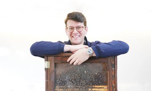 A smiling young man leans on a wooden observation beehive box, with bees visible through the glass panel.