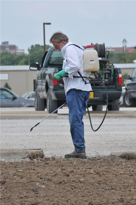 Man applying a spray pesticide while wearing personal protective equipment: gloves, boots, long pants, long sleeve shirt, and glasses
