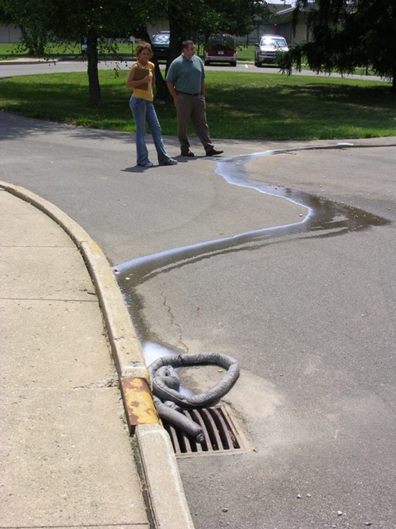 Two people watching a flow of liquid run towards and down a storm drain.