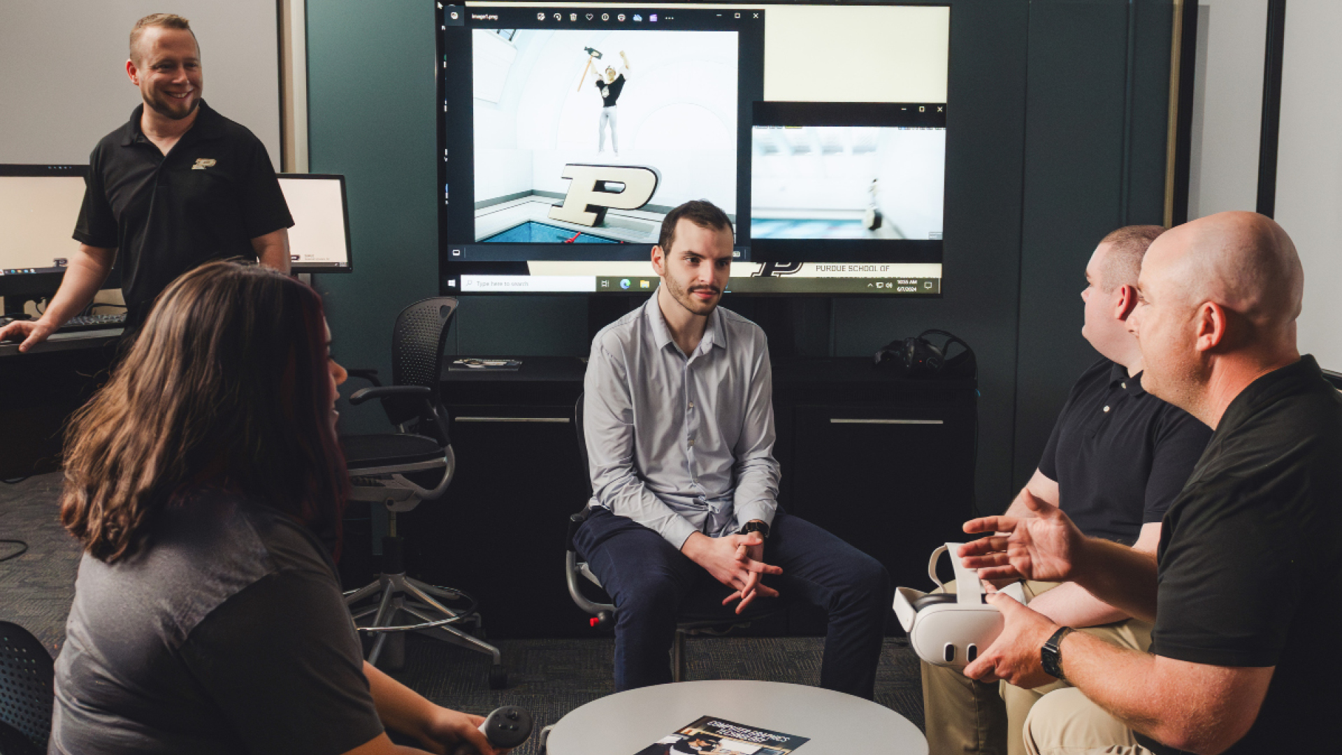 Three men and one woman seated, one man standing, in front of a Purdue Pete animation on a large screen