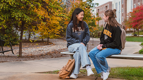 Two students sitting and talking on a bench on the Purdue campus during autumn.
