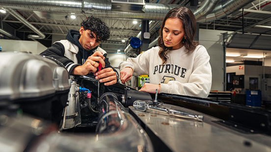 Two students working on a mechanical engine inside a workshop setting.