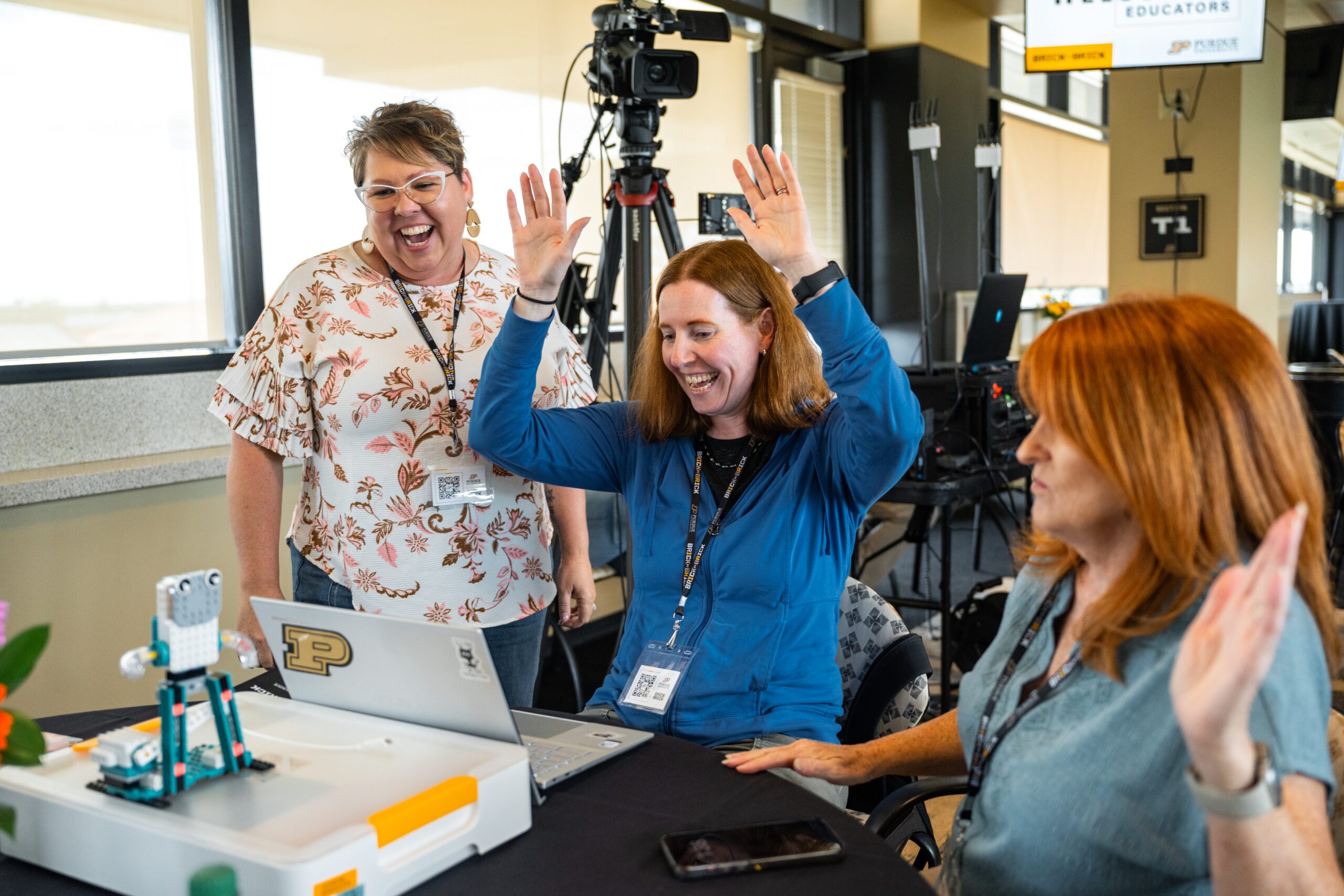Three individuals celebrate enthusiastically at a table with a robot and laptop, in a room equipped with cameras and production gear.