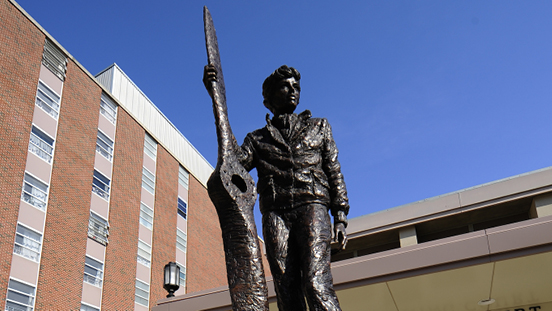 A bronze statue of Amelia Earhart holding an airplane propeller blade, situated outdoors in front of a brick multi-story building under a clear blue sky.