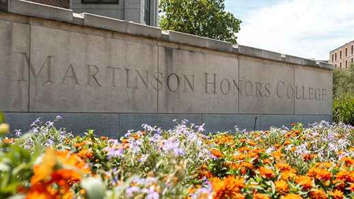 Stone signage reading John Martinson Honors College behind a bed of colorful flowers.