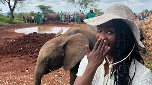 Woman in a sunhat holding her hand over her mouth while standing near a baby elephant in an outdoor reserve setting.