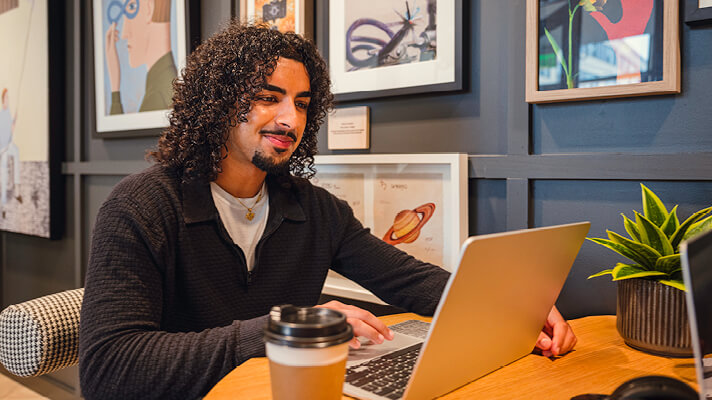 Student working on a laptop at a café table with a coffee cup beside him.