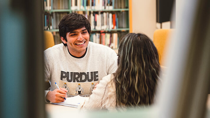 Student wearing a Purdue sweatshirt seated at a desk and smiling during a study session in the library.