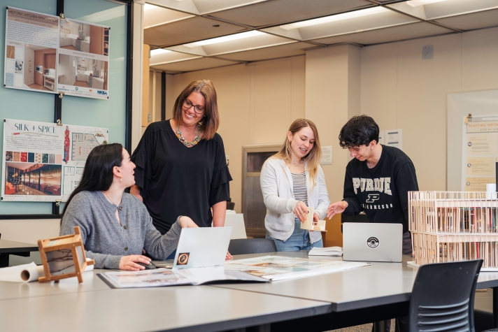 Students and an instructor gathered around a table in a classroom, working on laptops and reviewing architectural models and design boards displayed on the walls.