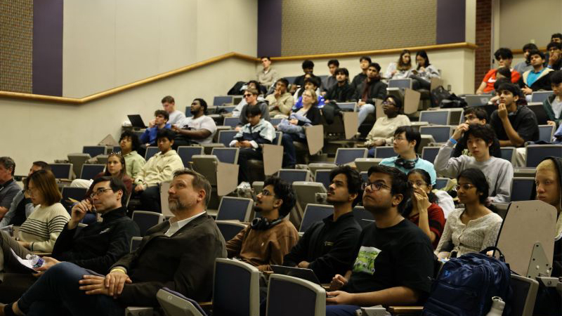 Hack Indy 2026 participants seated in a tiered lecture hall, listening and working on laptops during the event.