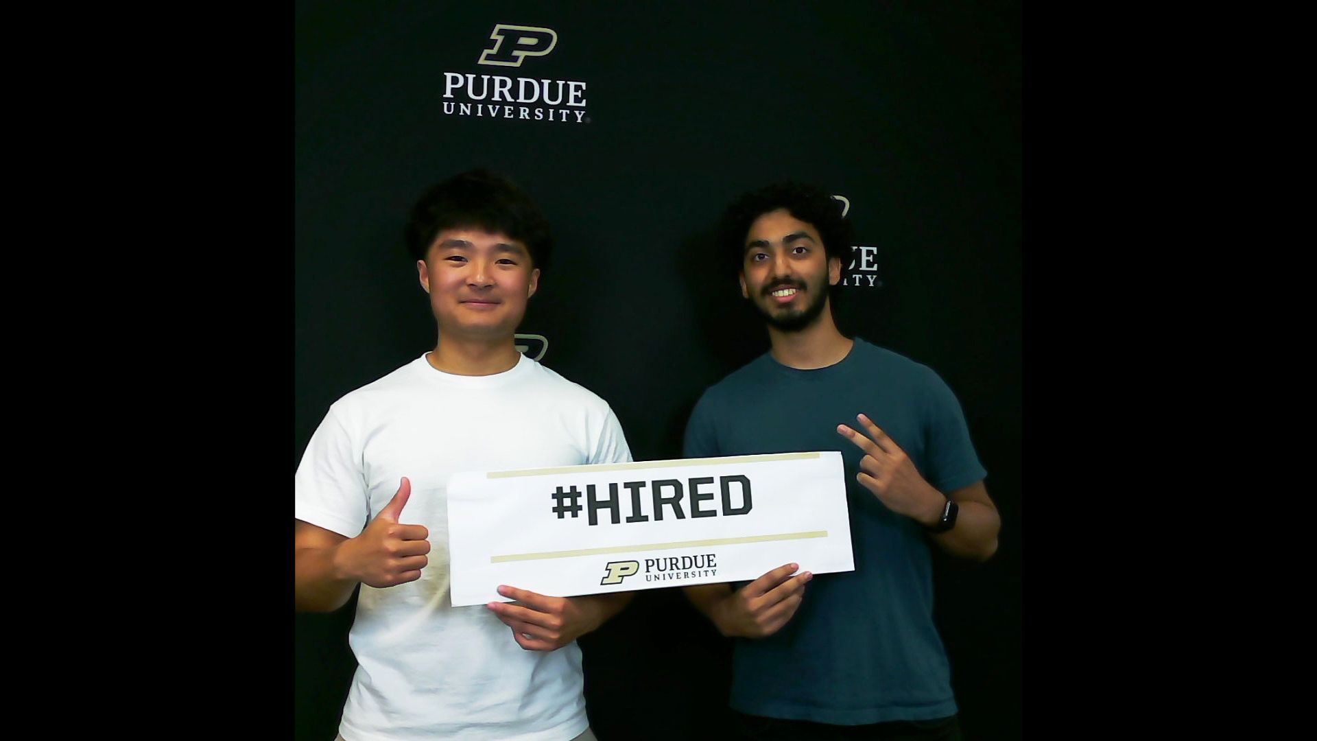 Two Purdue students pose together in front of a Purdue University backdrop holding a sign that reads #HIRED