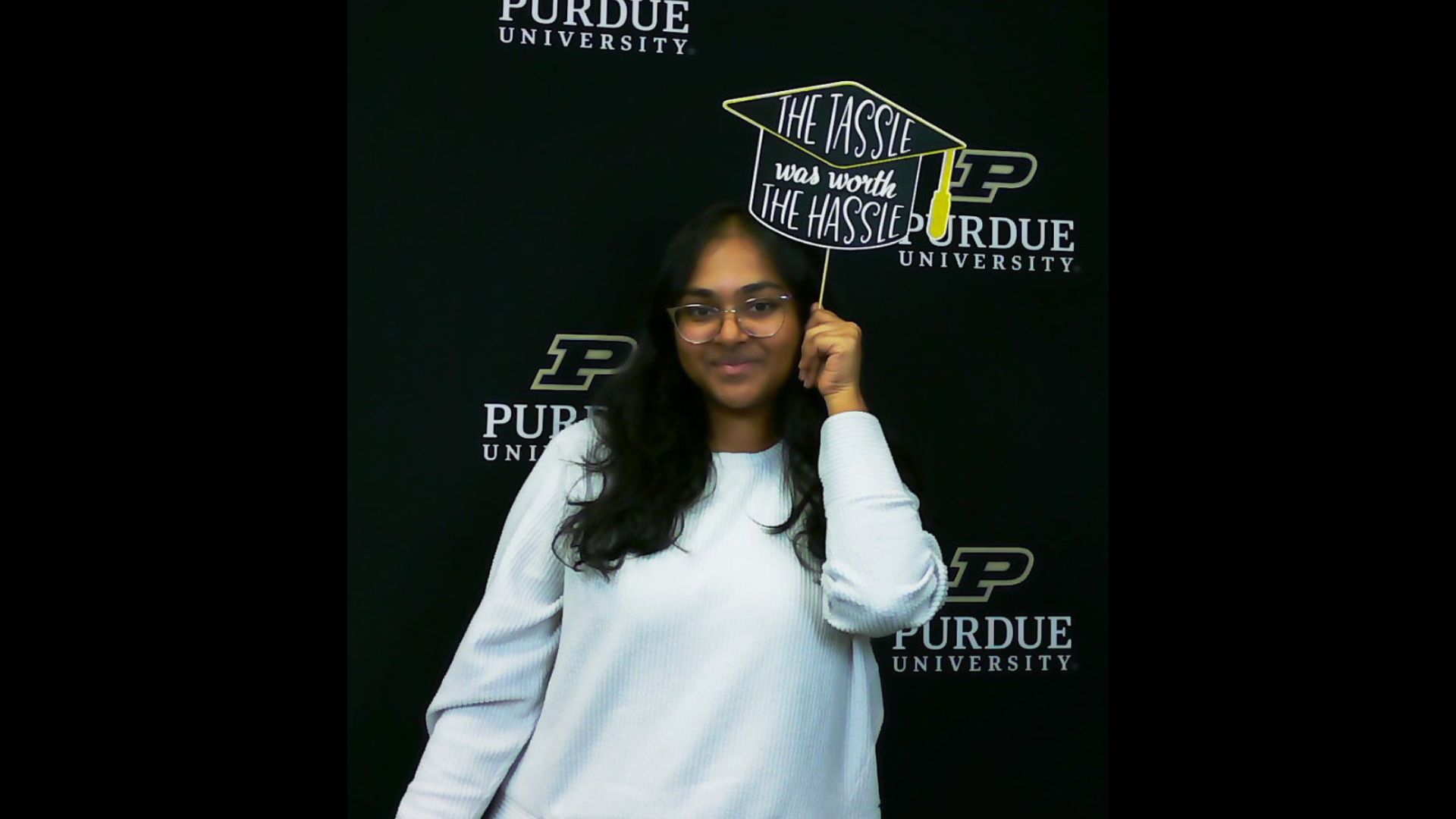 A Purdue student stands in front of a Purdue University backdrop holding a sign that reads “The tassel was worth the hassle.