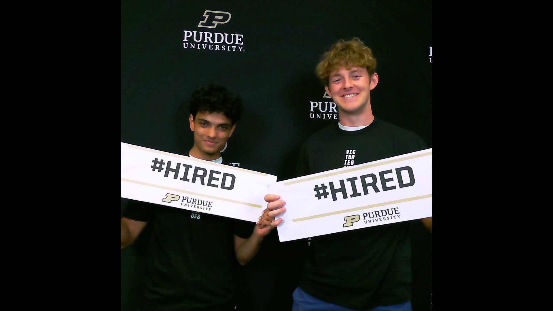 Two Purdue students stand in front of a Purdue University backdrop holding signs that read HIRED.