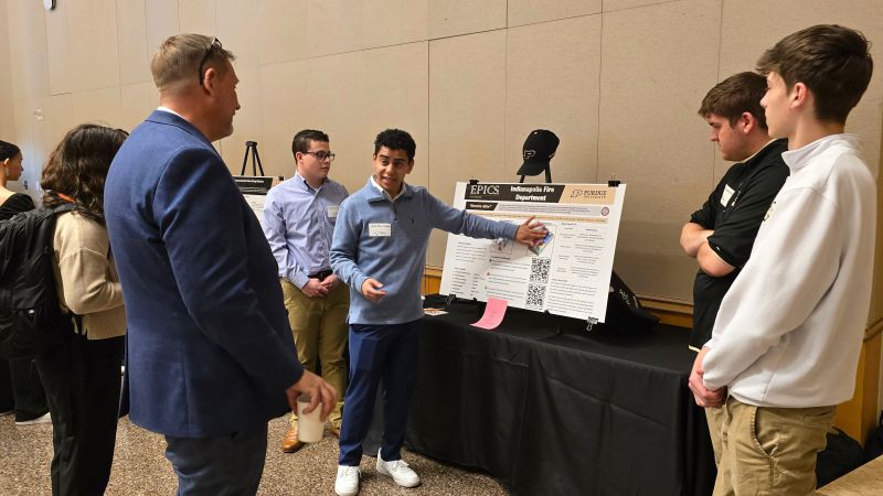 Attendees gather around a student project table at a Purdue event in Indianapolis, discussing an EPICS display