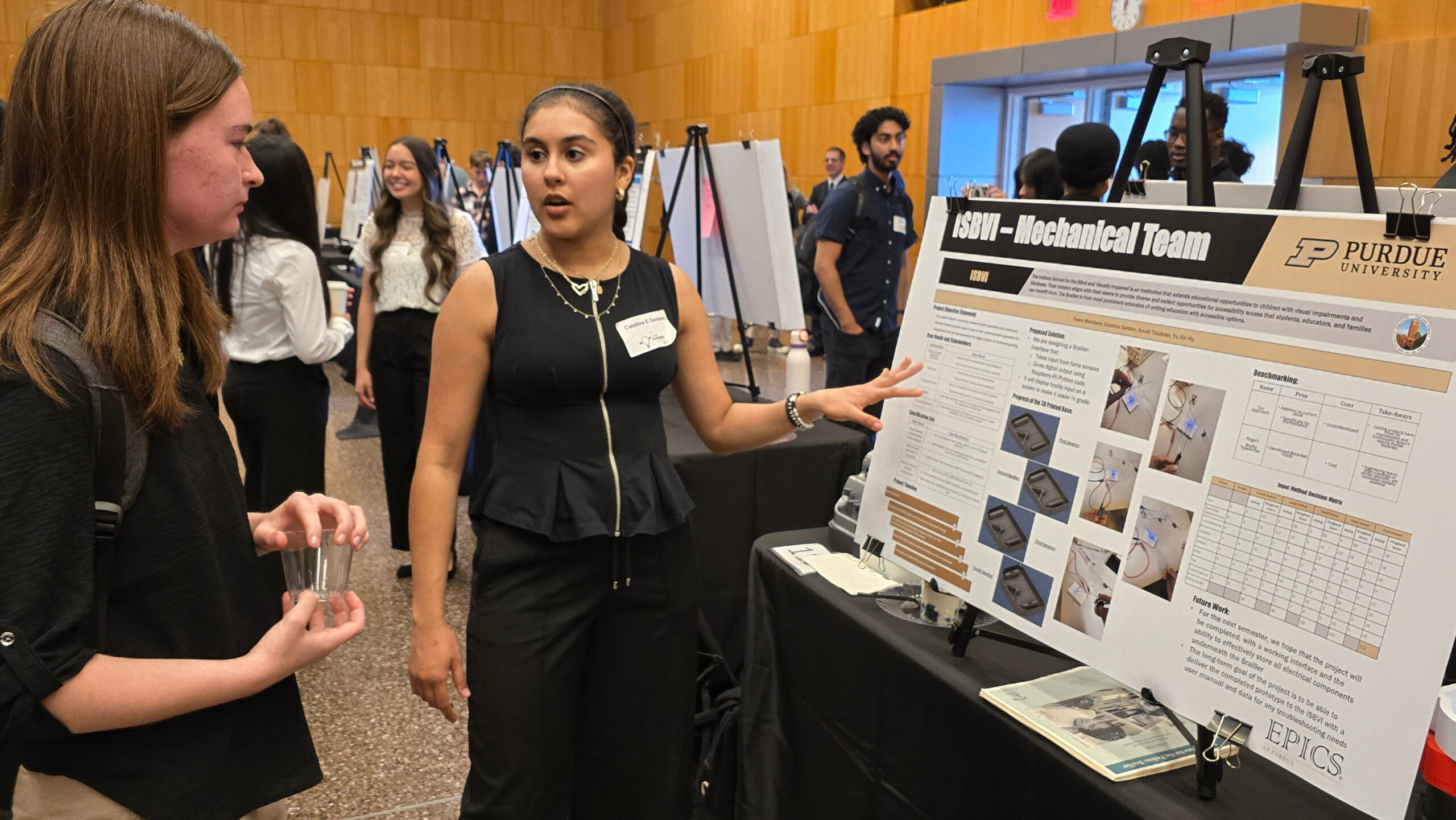 A student explains a mechanical team project poster during a Purdue event, with attendees reviewing diagrams and data