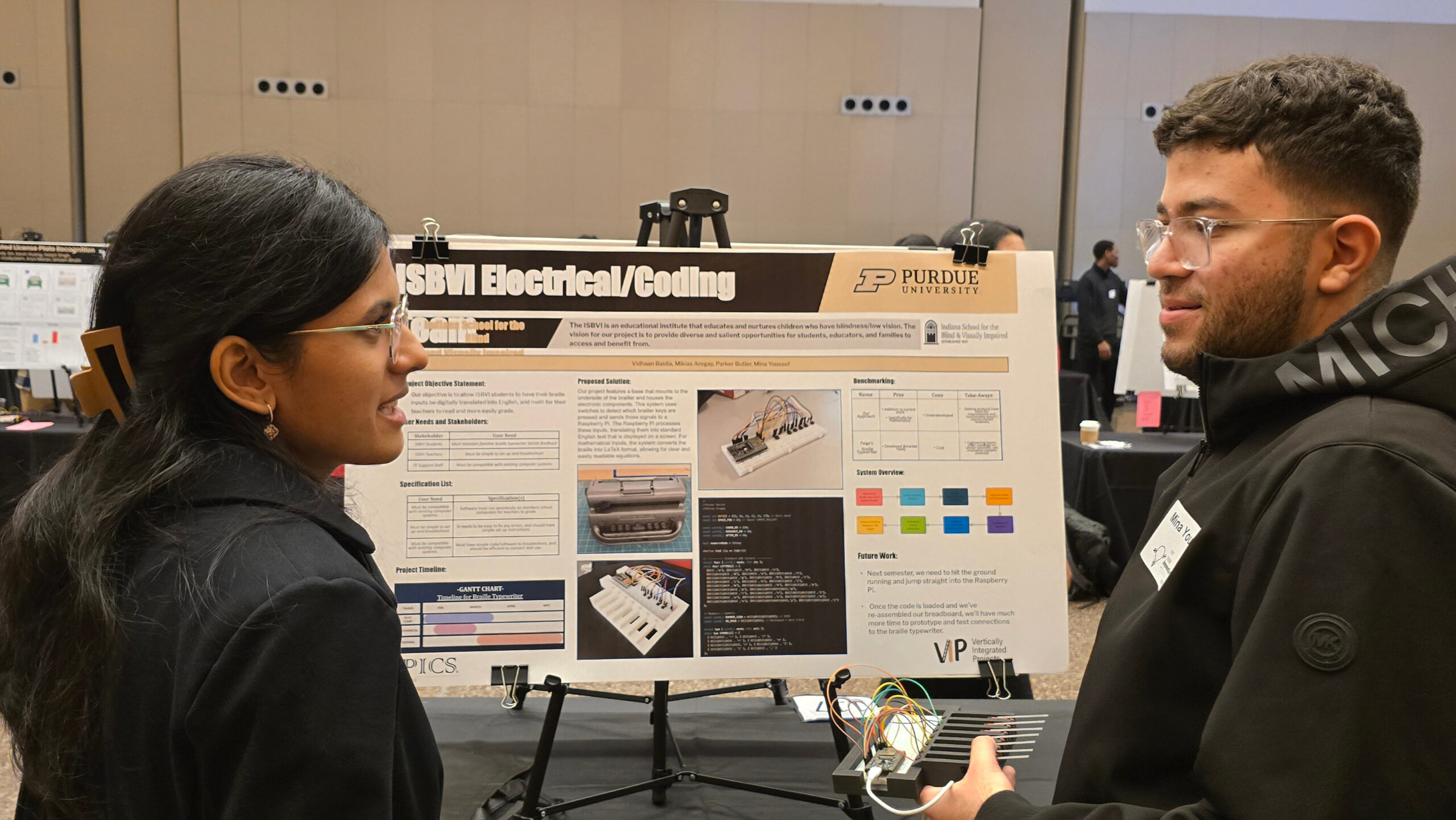 Students discuss an electrical and coding project at a Purdue showcase, standing beside a project poster and prototype wiring