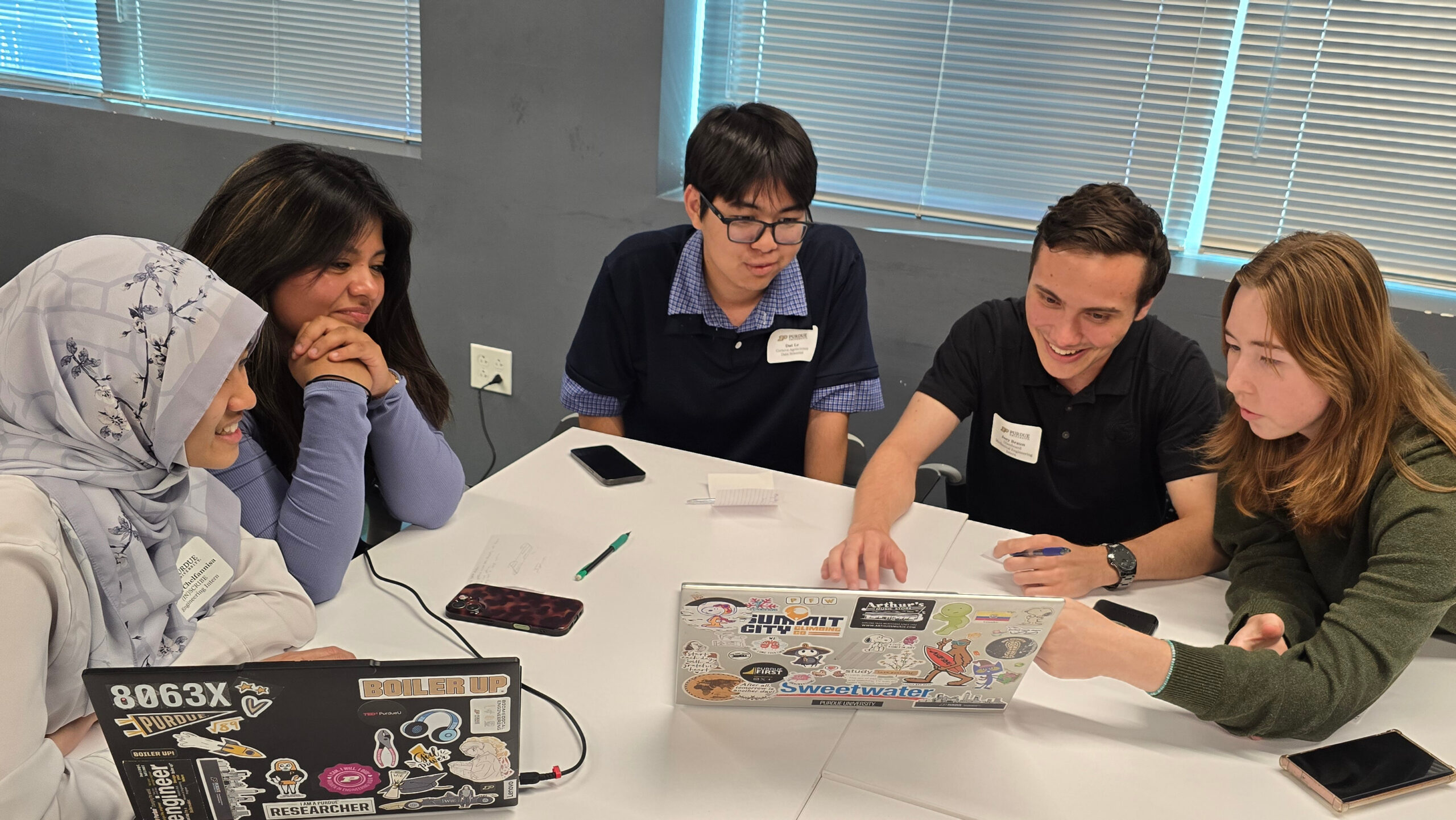 Five students sitting around a table in a room looking at a laptop