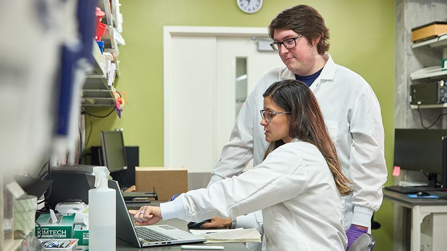 Scientists in white lab coats discussing in front of a laptop computer