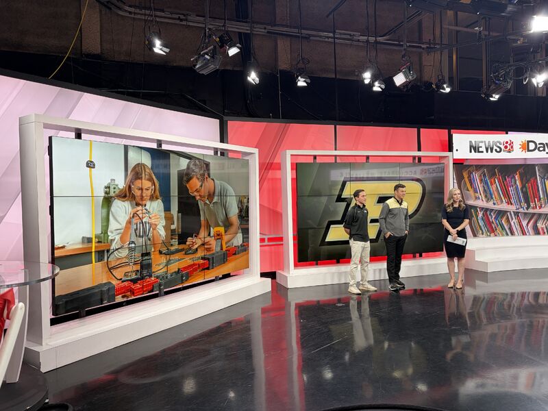 Students and anchors standing in the middle of a newsroom with the Purdue logo and image of students projected on the screen