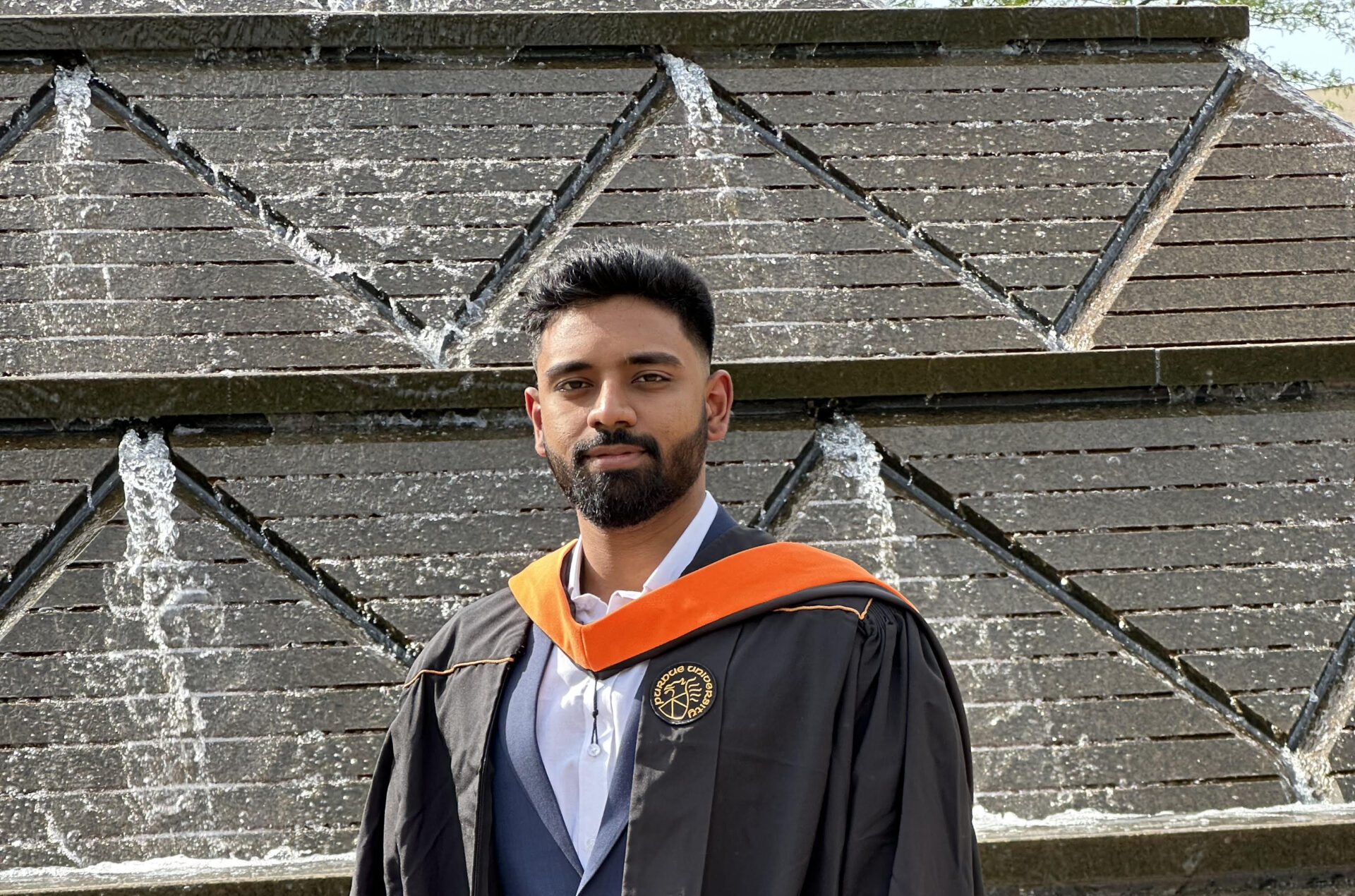 A student wearing a graduation gown and hood standing in front of a water structure