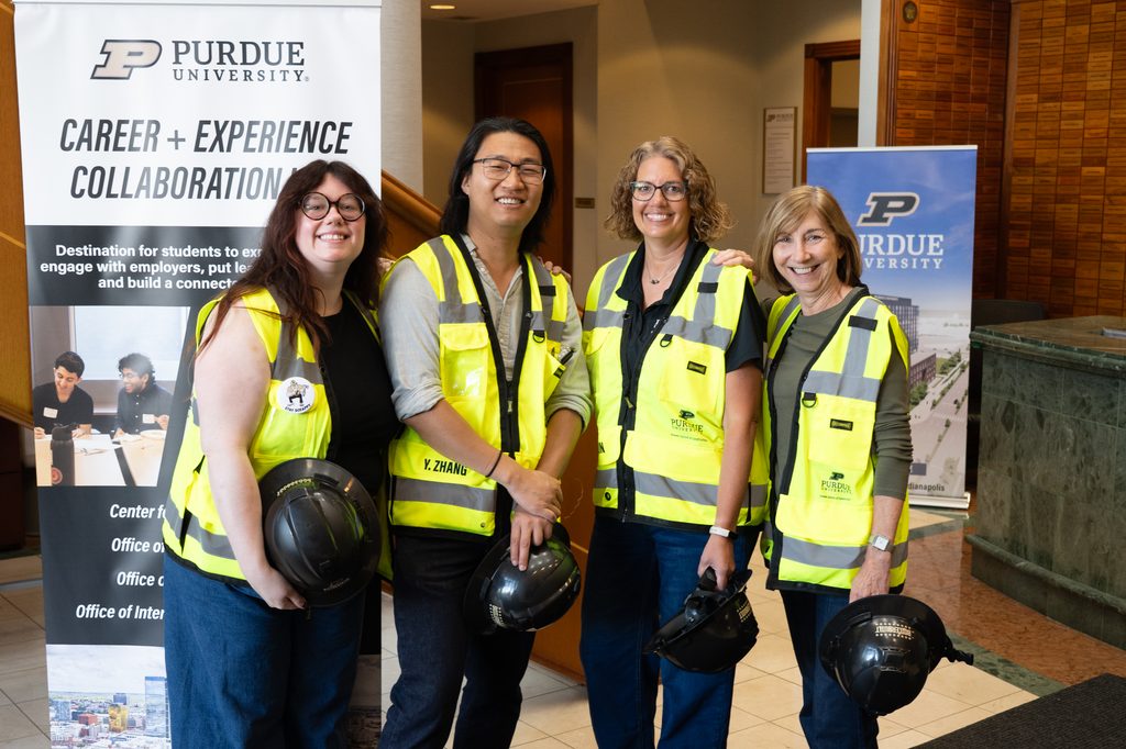 Four Purdue faculty and staff members wearing safety vests hold hard hats during a university event.