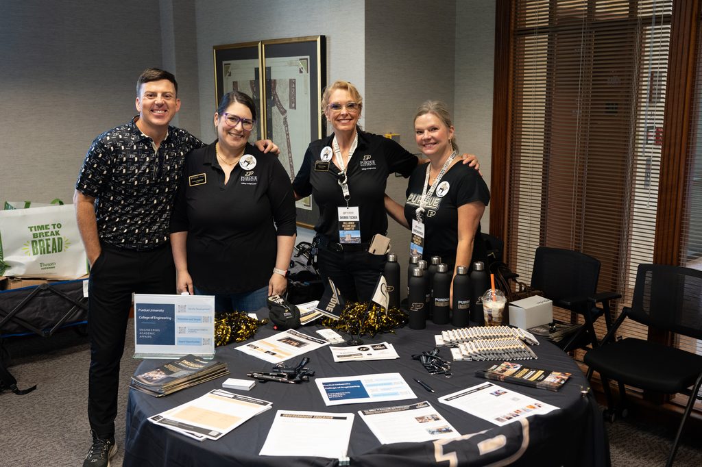 Four Purdue representatives stand behind a table with engineering program materials and giveaways.