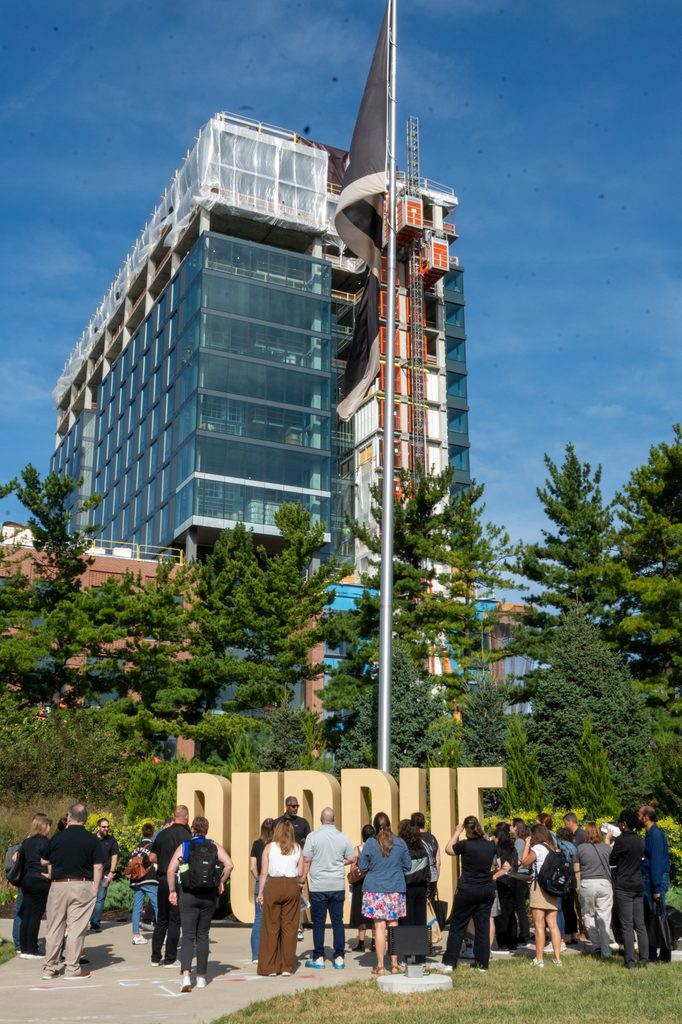 A group gathers near the gold letters spelling out Purdue in front of the Purdue flag in Indianapolis with the Academic Success Building in the background.