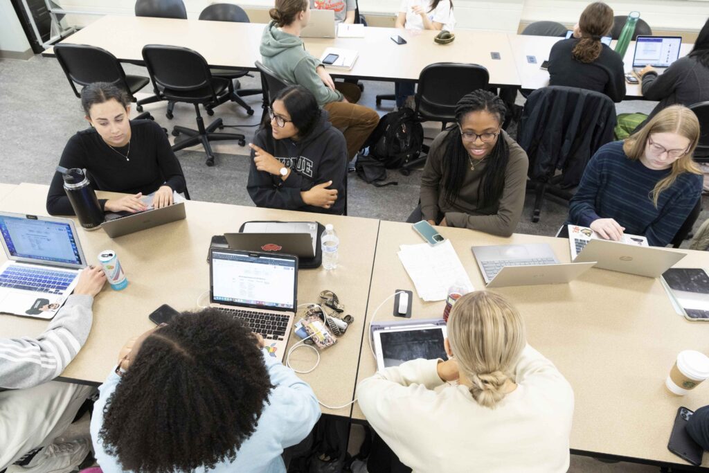 Students collaborate around tables in a classroom, with laptops and tablets open, some engaged in conversation and others focused on their work