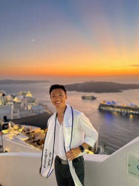 Alan Suen poses at a scenic overlook of a harbor with a vibrant sunset