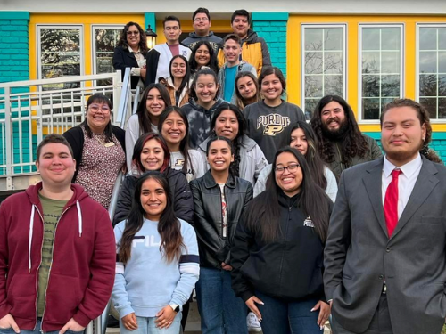 20 young Latinx and first-generation students standing in front of a building tha tis yellow, turquoise and pink