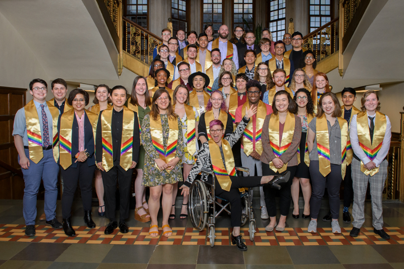 60 members of the Lavender Graduation Class of 2019; standing in group on grand staircase in Purdue Memorial Union. Each graduate is wearing a gold stole with rainbow fabric stitched onto it.