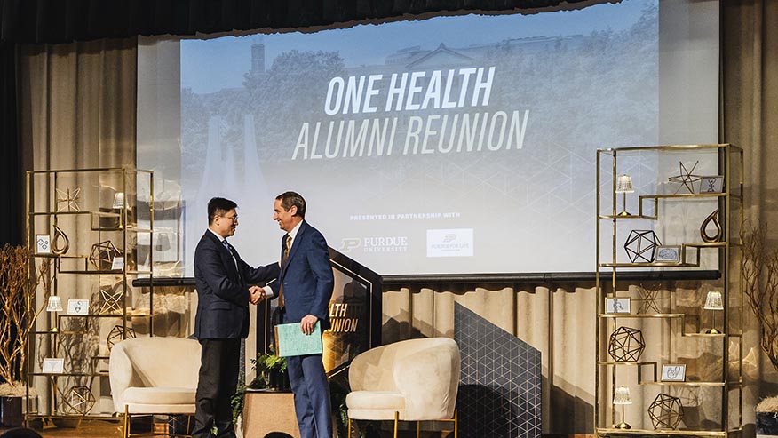 Two men shake hands in front of a projection screen that reads One Health Alumni Reunion.