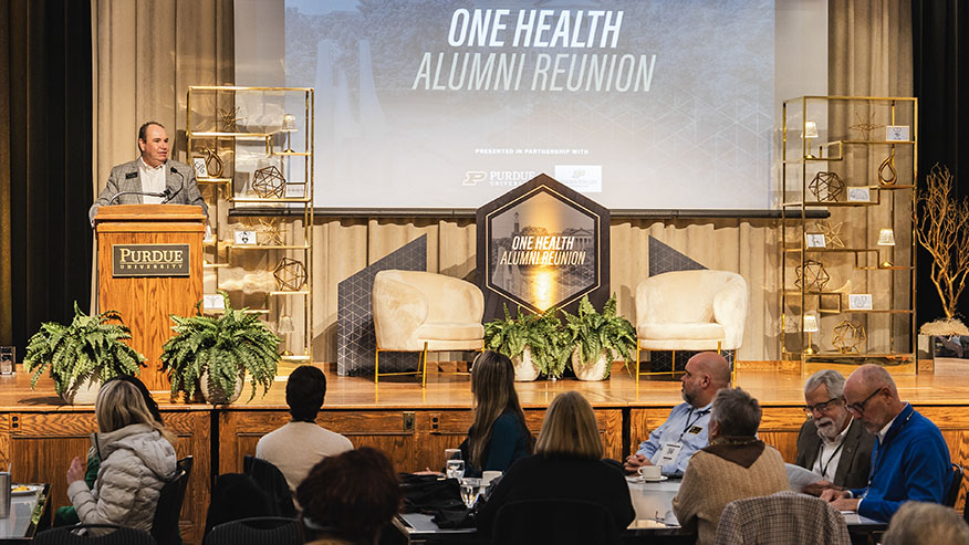 A man speaks in front of a projection screen that reads One Health Alumni Reunion.