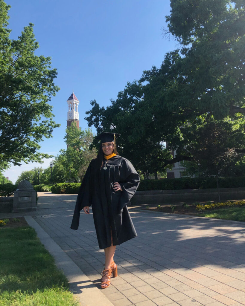 A woman in a cap and gown poses on a sidewalk with the Purdue Bell Tower in the distance.