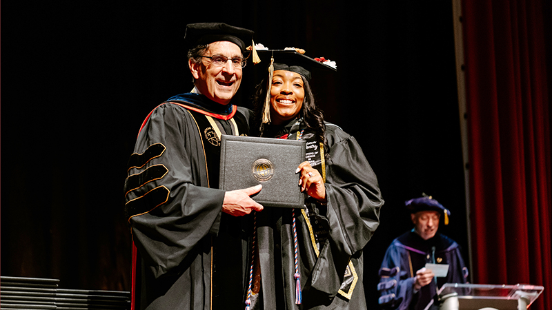 Purdue Global Chancellor Frank Dooley poses for a picture onstage with a graduating student accepting a diploma.