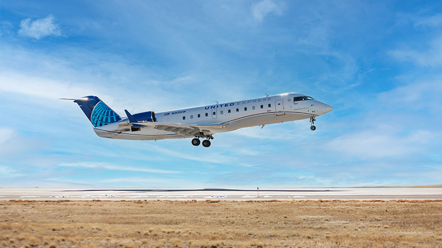 A United Express CRJ-200 regional jet with landing gear extended takes off from a runway, flying above a dry, grassy landscape under a bright blue sky.