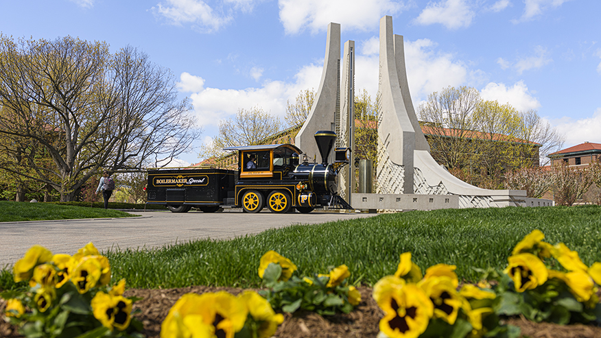 The Boilermaker Special, Purdue University’s official mascot train, parked in front of the Class of 1939 Water Sculpture on a sunny spring day with yellow flowers blooming in the foreground.