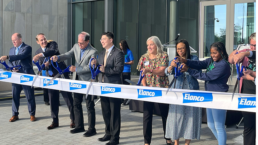 President Mung Chiang and others are standing in a line, each holding a pair of scissors and cutting a long white ribbon with the word 'Elanco' printed on it. This event is a ribbon-cutting ceremony taking place near downtown Indianapolis.