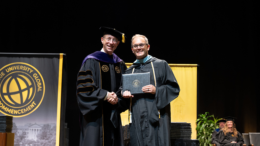 Pat Doney and Jon Harbor shaking hands and holding a diploma on a stage at commencement