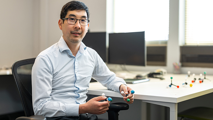 A man wearing glasses holds small models of molecules while sitting in a desk chair next to a table.