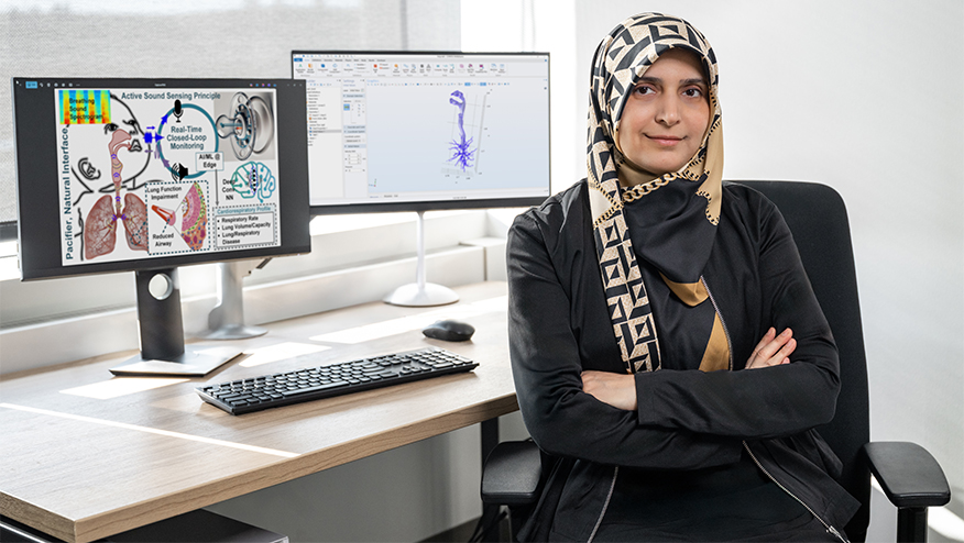 A woman sits with her arms crossed in front of two computer screens showing her research