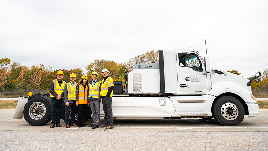 Five Purdue professors wearing hard hats and reflective vests stand in front of an electric semitractor on a roadway