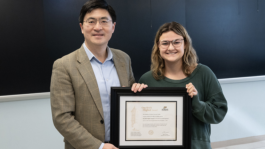 Two people holding a framed award certificate with a chalkboard in the background.
