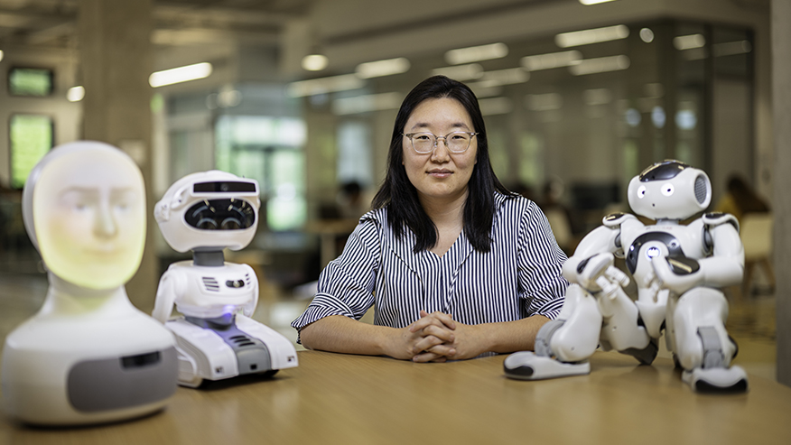 A smiling scientist sits at a desk with three vaguely humanoid robots.