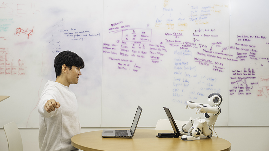 A human and a robot sit across from each other at a desk, both engaging in a stretching exercise.