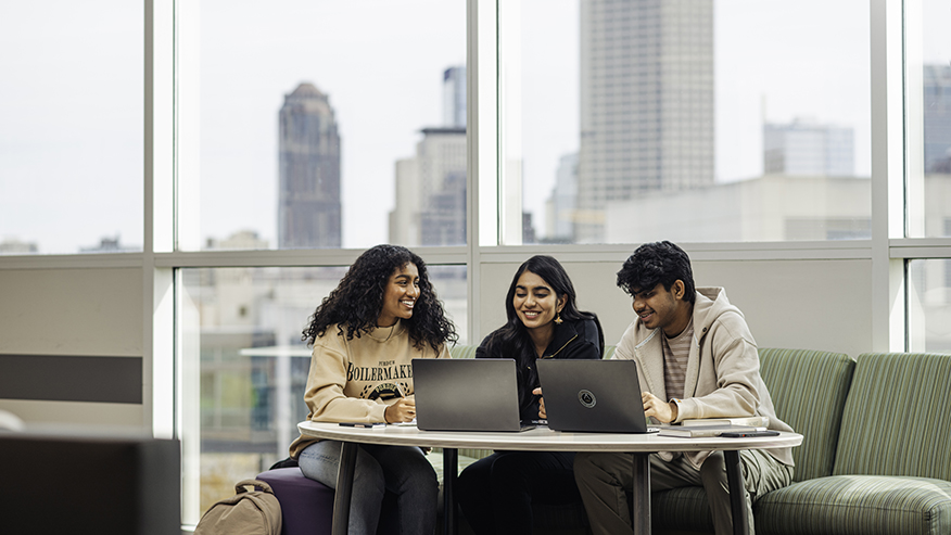 Three Purdue students sitting at a table with two laptops open; the Indianapolis skyline is in the background