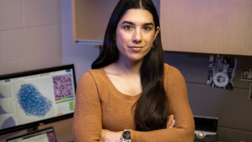 A woman with long black hair stands, arms crossed, before a computer screen on a table.